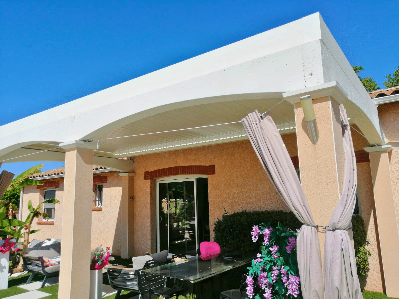 Pergola blanche cintrée sur terrasse aménagée d'une maison ocre avec mobilier de jardin.