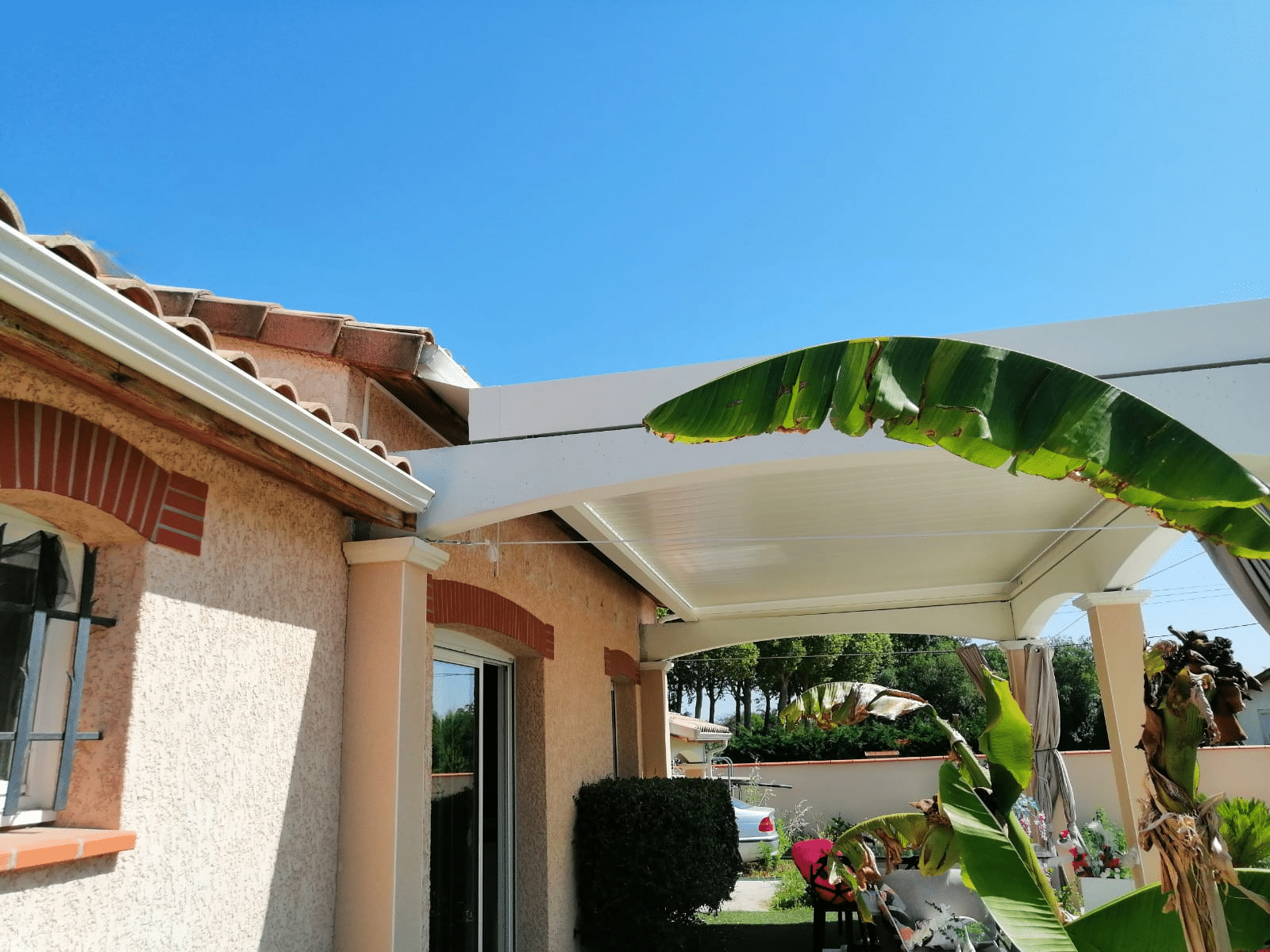 Pergola blanche moderne sur terrasse avec bananier et toit en tuiles méditerranéen.