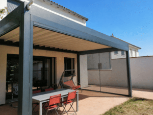 Pergola bioclimatique grise anthracite et toile rétractable et moustiquaires latérales sur une terrasse avec table et chaises rouges.