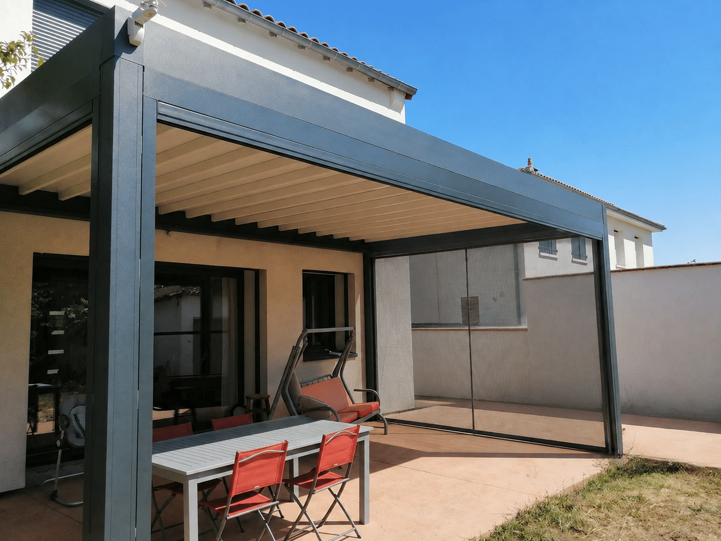 Pergola bioclimatique grise anthracite et toile rétractable et moustiquaires latérales sur une terrasse avec table et chaises rouges.