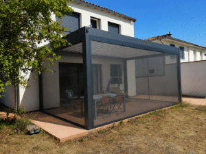 Pergola bioclimatique grise anthracite et toile rétractable et moustiquaires latérales sur une terrasse avec table et chaises rouges.