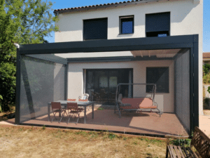 Pergola bioclimatique grise anthracite et toile rétractable et moustiquaires latérales sur une terrasse avec table et chaises rouges.